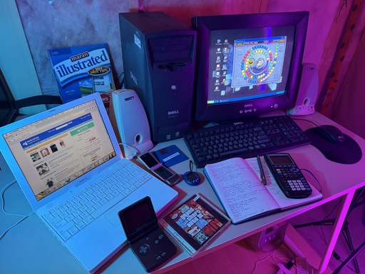 A staged early-2000s computer desk setup lit with blue and magenta accent lighting. On the left, a white iBook G4 laptop displays SpaceHey, a modern MySpace-style social network, in a Firefox-based browser. In the center is a Dell Dimension 2200 tower connected to a Dell CRT monitor showing Windows XP with the game Zuma Deluxe running, using a Dell keyboard and mouse. Beige Harman/Kardon desktop speakers sit beside the monitor. Spread across the desk are period accessories including a Motorola Razr flip phone, a blue Tamagotchi Connection, a TI-84 graphing calculator resting on an open handwritten notepad, a black Game Boy Advance SP, and a blue 3.5-inch floppy disk labeled “Plop Bootloader.” A GTA III jewel case lies near the front edge of the desk, and a Maran Illustrated Windows XP: 101 Tips book stands behind the computers.